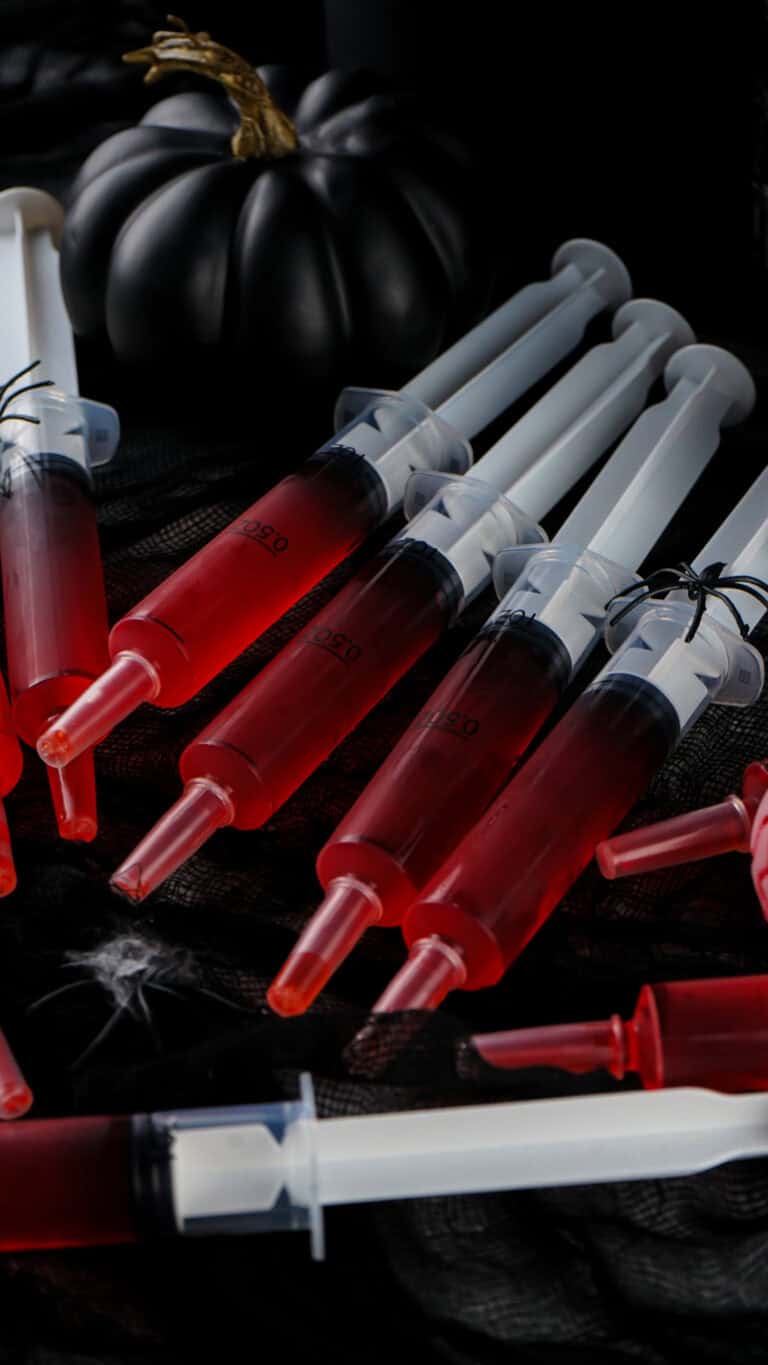 Several syringes filled with red liquid are displayed on a dark surface, resembling eerie Syringe Jello Shots, accompanied by small decorative spiders and a black pumpkin in the background.