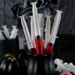 A collection of syringe jello shots filled with red liquid displayed in a black cup, surrounded by additional syringes, black roses, a black pumpkin, and a decorative eyeball on the table.