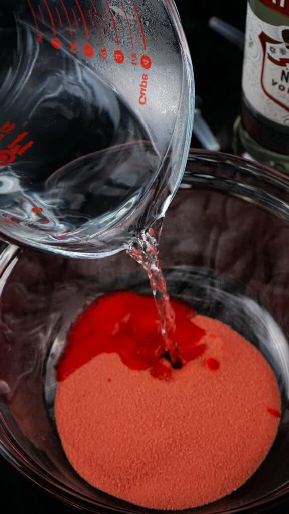 A measuring cup pours clear liquid into a glass bowl containing red powder, prepping the mixture for vibrant Jello Shots.