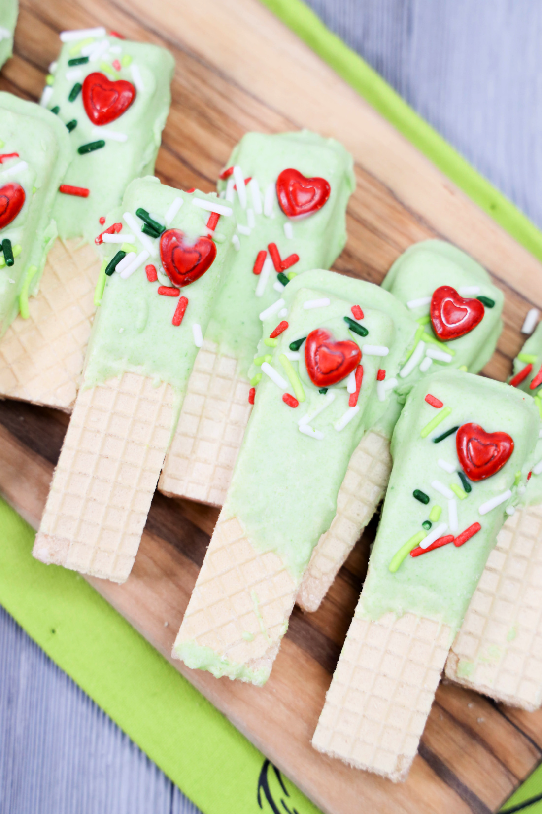 Grinch Dipped Wafers: Green wafer treats topped with red heart candies and colorful sprinkles, arranged on a wooden board.