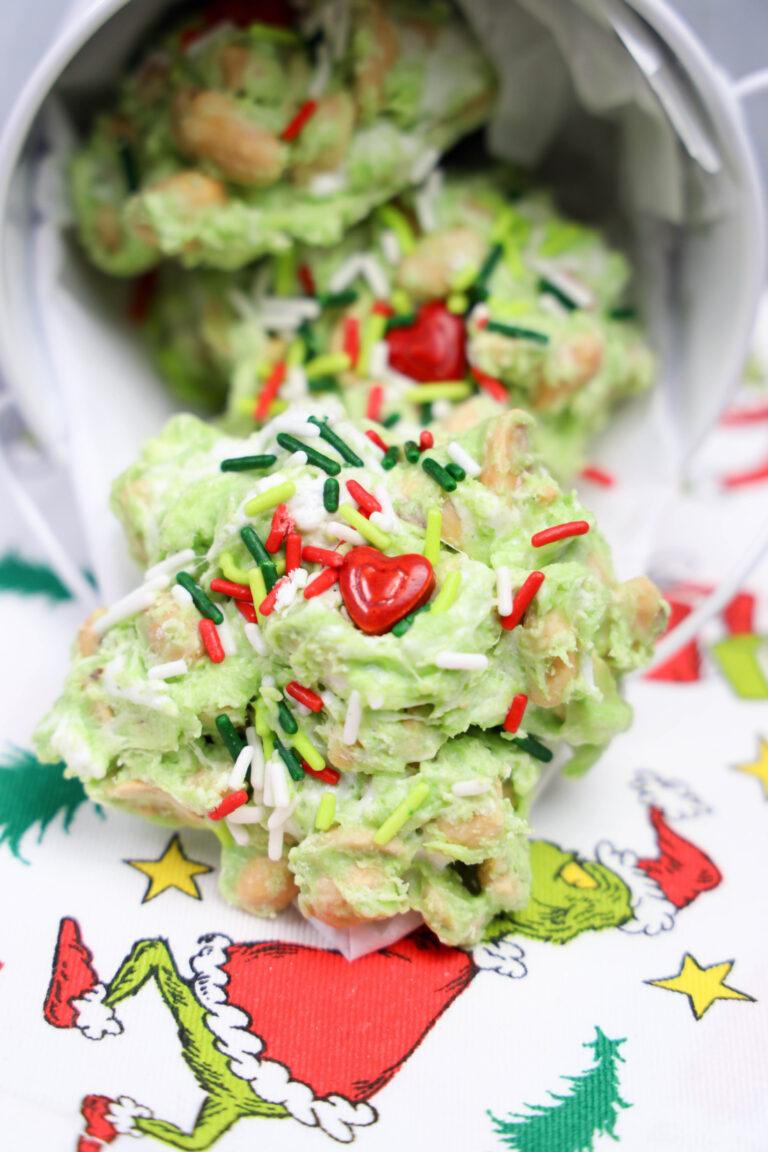 A festive snack mix of Grinch Poop Peanut Clusters with green coating, colorful sprinkles, and heart-shaped decorations spills from a container onto a holiday-themed cloth.