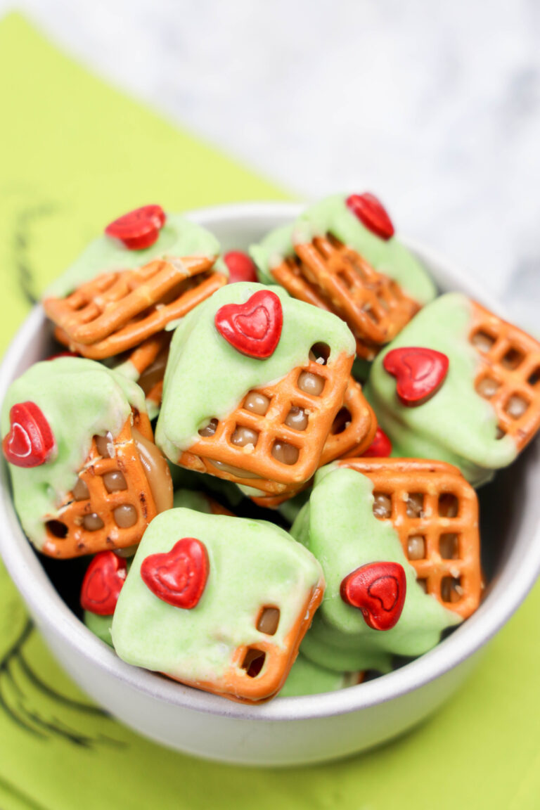 A bowl of Grinch Heart Pretzel Bites features square pretzels coated in green frosting, each topped with a red heart-shaped candy.