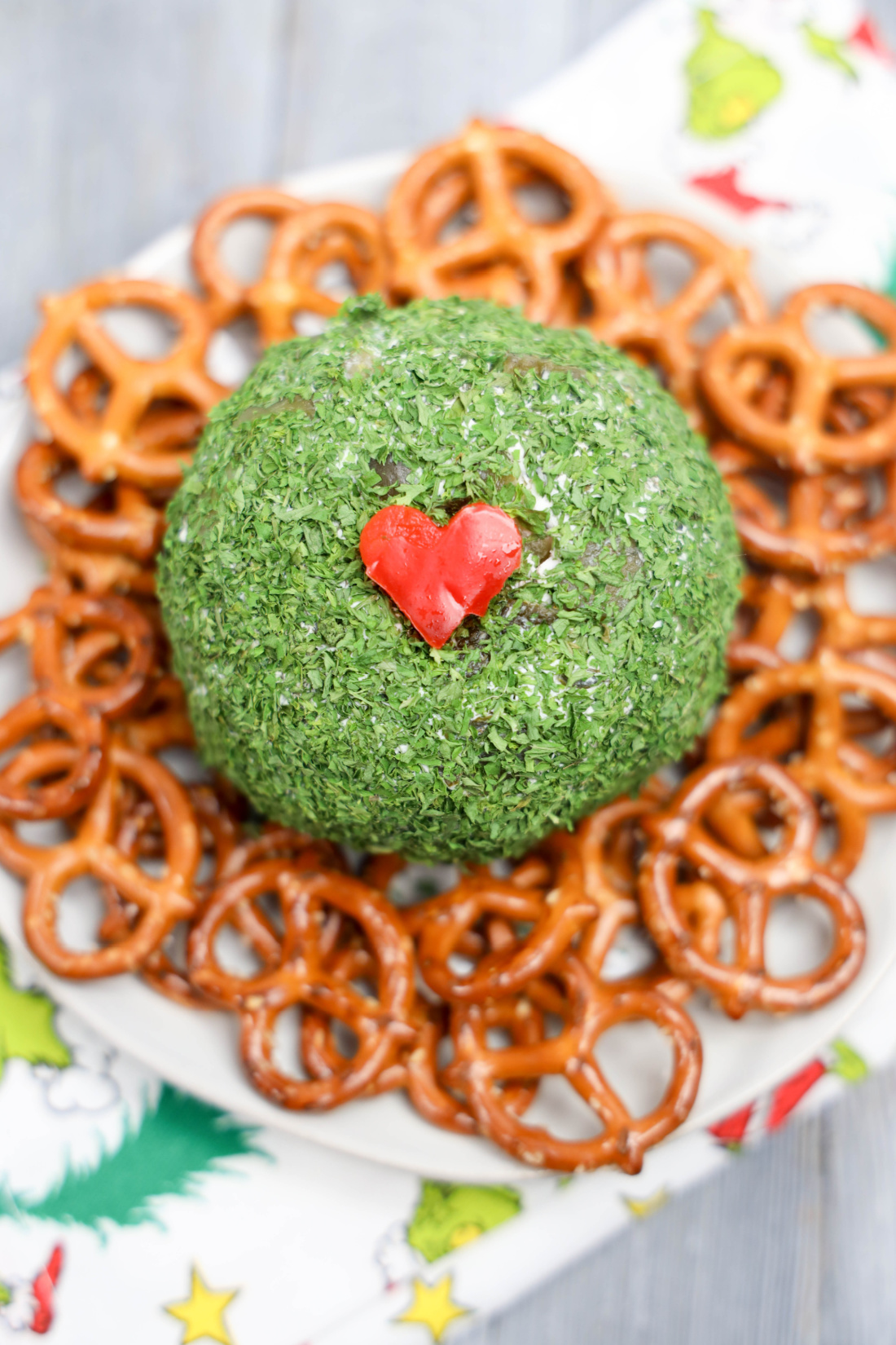 The Grinch Cheese Ball, coated with green herbs and topped with a red heart-shaped decoration, is surrounded by pretzels on a white plate.