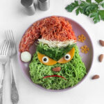 A whimsical plate of Grinch-themed vermicelli pasta features red and green noodles crafted into a festive face. Cucumber and yellow pepper form the eyes, topped with dill. Nearby, a fork, spoon, and sprig of parsley complete this holiday character masterpiece.