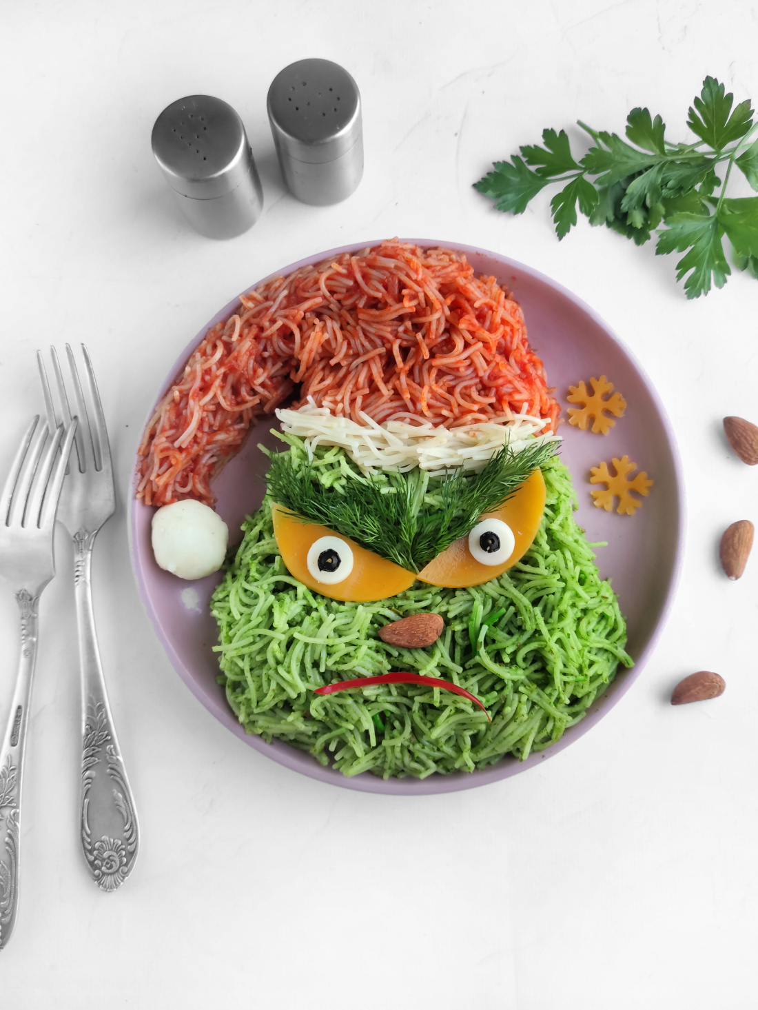 A whimsical plate of Grinch-themed vermicelli pasta features red and green noodles crafted into a festive face. Cucumber and yellow pepper form the eyes, topped with dill. Nearby, a fork, spoon, and sprig of parsley complete this holiday character masterpiece.