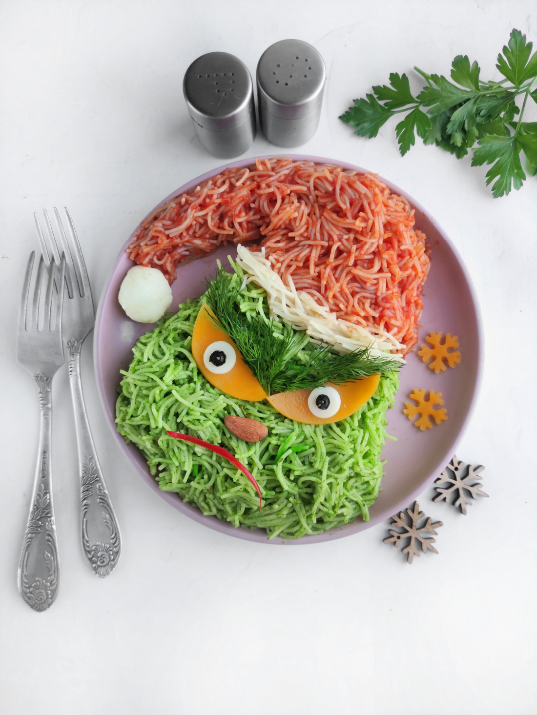 A creative dish arranged to resemble the Grinch's face, featuring vermicelli pasta in green and red hues, accompanied by vibrant vegetables and fresh herbs. This playful plate is completed with utensils and seasoning containers nearby for a festive touch.