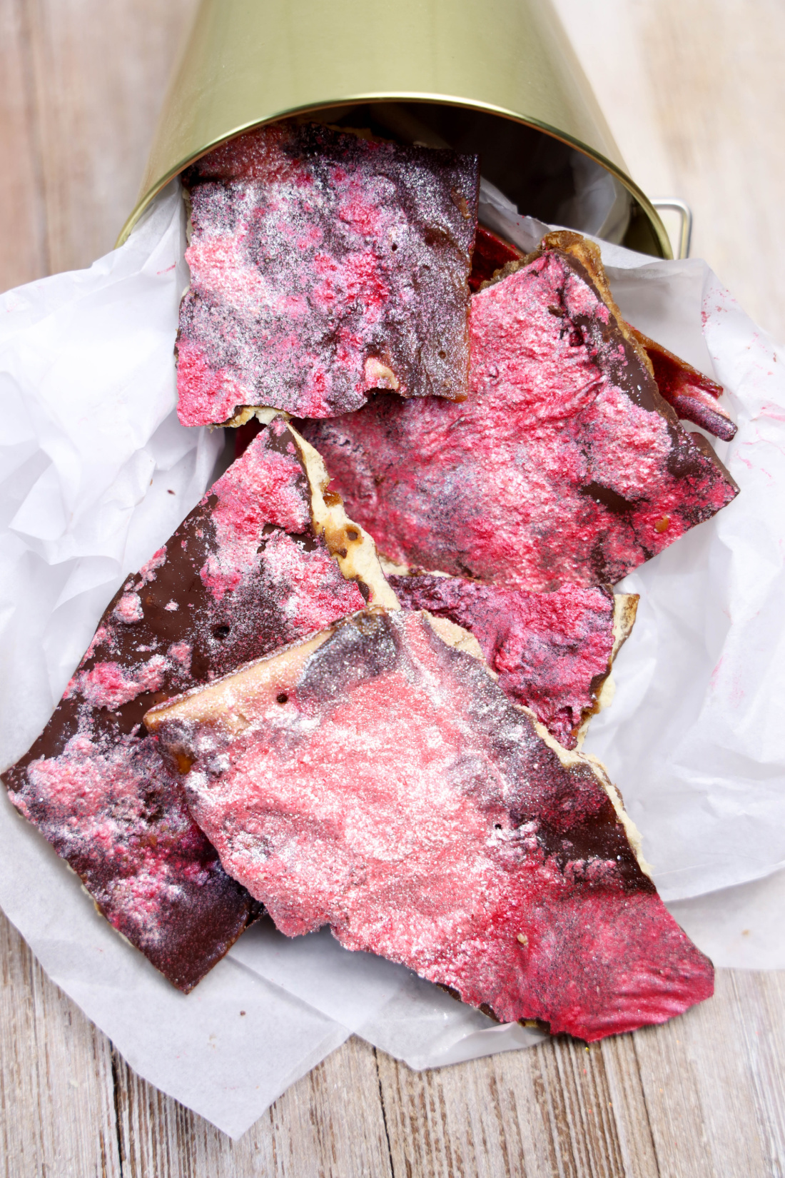 Crimson-colored chocolate bark with a dusting of white sprinkles, reminiscent of Glitter Valentine's Cracker Candy, partially spills out of a metal container onto white parchment paper.