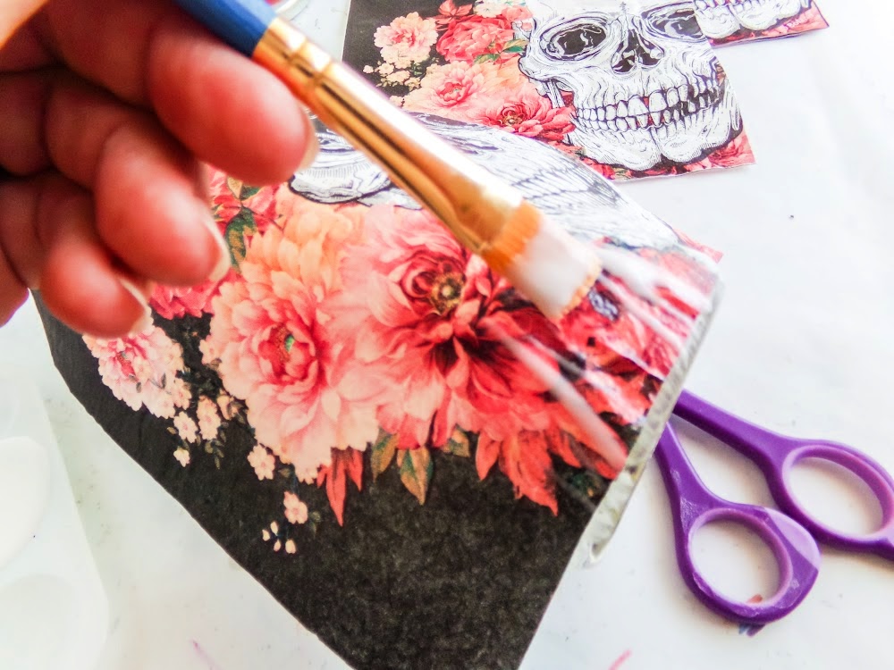 Skull Cookie Mix in a Jar 7 A hand applies glue with a brush to paper featuring skull and flower designs. Scissors are visible in the background.