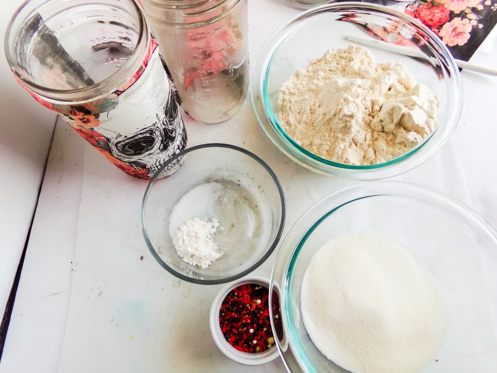 Skull Cookie Mix in a Jar 4 A table with glass bowls containing flour, sugar, a small amount of baking powder, and colorful sprinkles. Mason jars, one with a floral skull design, are in the background.