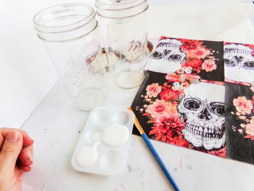 Skull Cookie Mix in a Jar 3 Two empty glass jars, a hand holding a brush, a paint palette with white paint, and a napkin featuring skulls and flowers on a table.