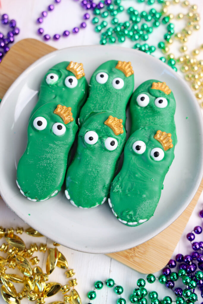 Green alligator-shaped cookies with candy eyes rest on a white plate, capturing the spirit of Mardi Gras Alligator Cookies. They are surrounded by colorful beads and bells.