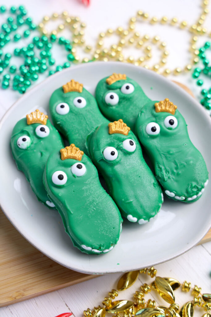 A plate of six enchanting Mardi Gras Alligator Cookies, each with googly eyes and tiny golden crown decorations, surrounded by festive beads.