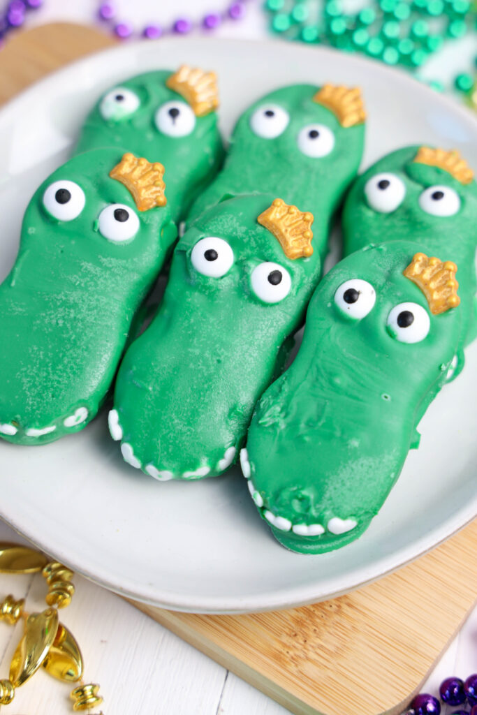 Mardi Gras Alligator Cookies with candy eyes and crown-shaped decorations rest on a white plate, surrounded by decorative beads.