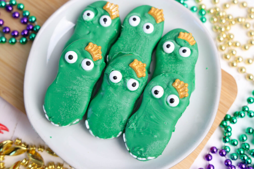 Plate with six green alligator cookies featuring googly eyes and golden scales, surrounded by colorful beads on a wooden surface. These Mardi Gras Alligator Cookies bring a playful touch to any celebration.