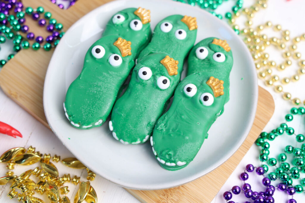 Mardi Gras Alligator Cookies in dinosaur shapes are artfully arranged on a white plate, surrounded by vibrant, colorful beads.