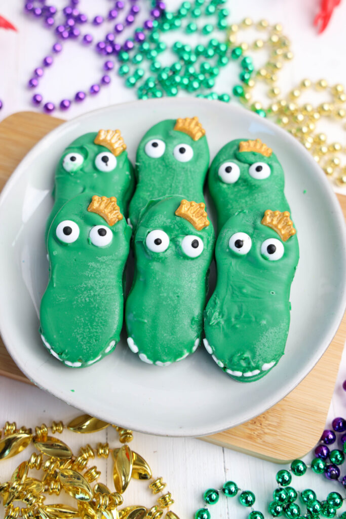 A plate of vibrant green alligator cookies, whimsically shaped like crocodiles with white eyes and shimmering gold details, surrounded by a festive array of colorful beads. Perfect for adding a playful touch to your Mardi Gras celebrations!.