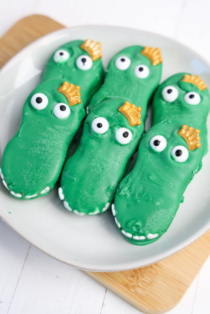 Green crocodile-shaped cookies, adorned with white and black eyes and yellow crowns, rest on a white plate. These delightful treats, resembling Mardi Gras Alligator Cookies, add a festive twist to any table setting.
