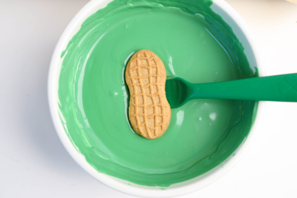 A peanut-shaped cookie on a green spoon rests in a bowl of vibrant green frosting, showcasing the whimsical charm of Mardi Gras Alligator Cookies.