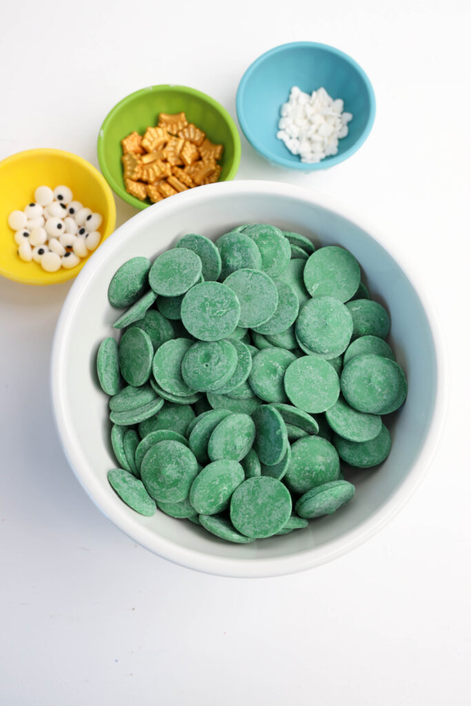 Green candy melts in a white bowl, surrounded by small bowls with white candy eyes, gold pieces, and snowflake sprinkles—perfect for crafting Mardi Gras Alligator Cookies.