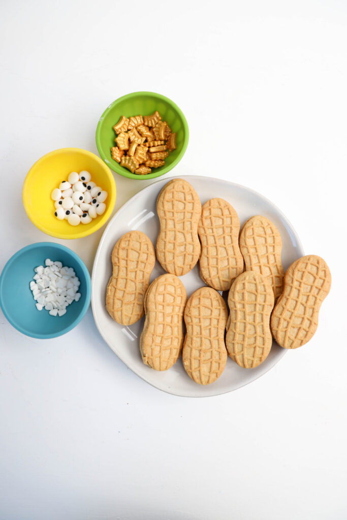 A plate of alligator-shaped cookies is surrounded by colorful bowls containing decorative candy eyes, snowflake sprinkles, and gold cereal pieces on a white background.