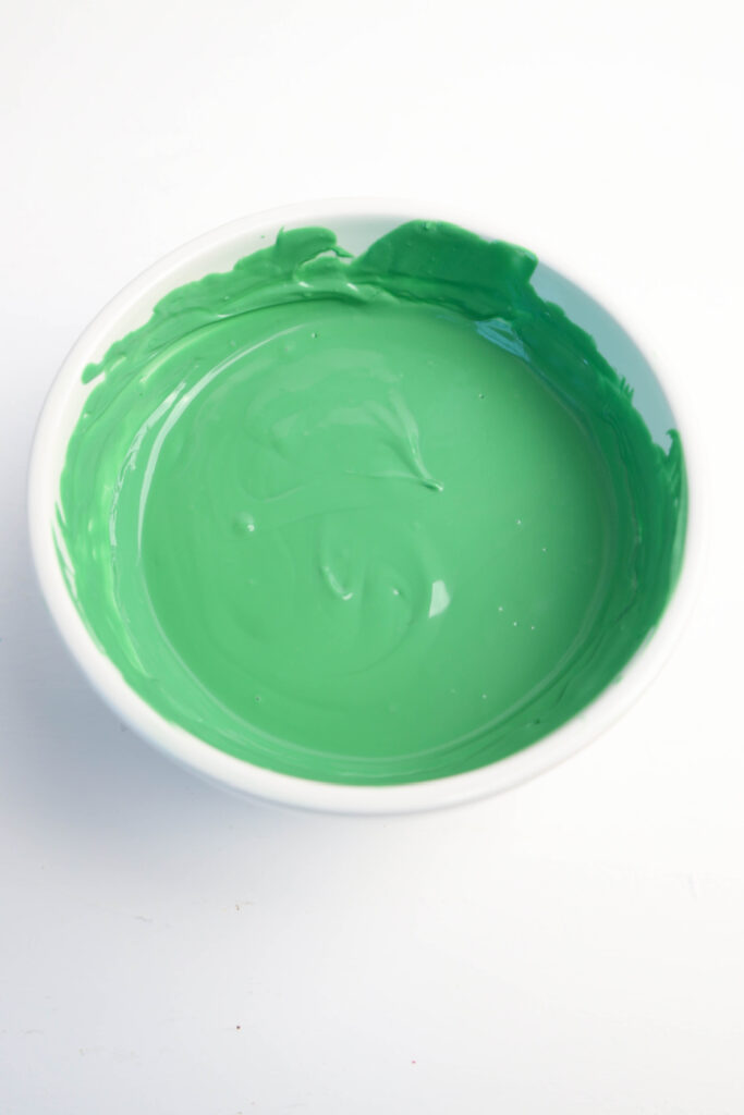 A white bowl filled with smooth, green paint sits on a white surface, reminiscent of the vibrant hues used to decorate festive Mardi Gras Alligator Cookies.