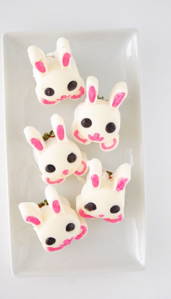 Chocolate Covered Strawberry Bunnies - Easy Easter Dessert 11 Five white chocolate treats shaped like bunny heads with pink ears, eyes, and noses are arranged on a white rectangular plate. These delightful confections bring to mind the charm of Chocolate Covered Strawberry Bunnies with their playful pastel touches.