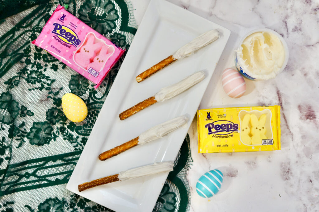 Peeps Pretzel Rods 4 Four Peeps Pretzel Rods coated in white chocolate are elegantly arranged on a rectangular white plate. Surrounding the display are packages of colorful Peeps marshmallows, vibrant decorative eggs, and a tempting tub of creamy white frosting.