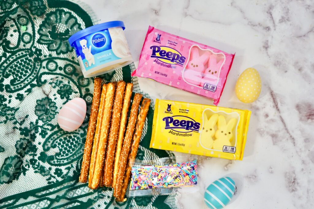 Peeps Pretzel Rods 3 Vanilla frosting, pretzel rods, multicolor sprinkles, yellow and pink Peeps marshmallows, and decorated eggs rest on a green lace cloth atop a marble surface.