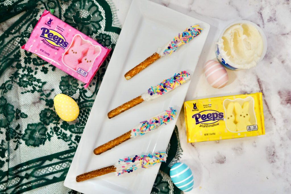 Peeps Pretzel Rods 5 Four decorated Peeps Pretzel Rods with white chocolate and vibrant sprinkles rest on a rectangular plate. Surrounding them are cheerful pink and yellow Peeps packages, a bowl of frosting, and pastel eggs, creating a delightful Easter-themed display.