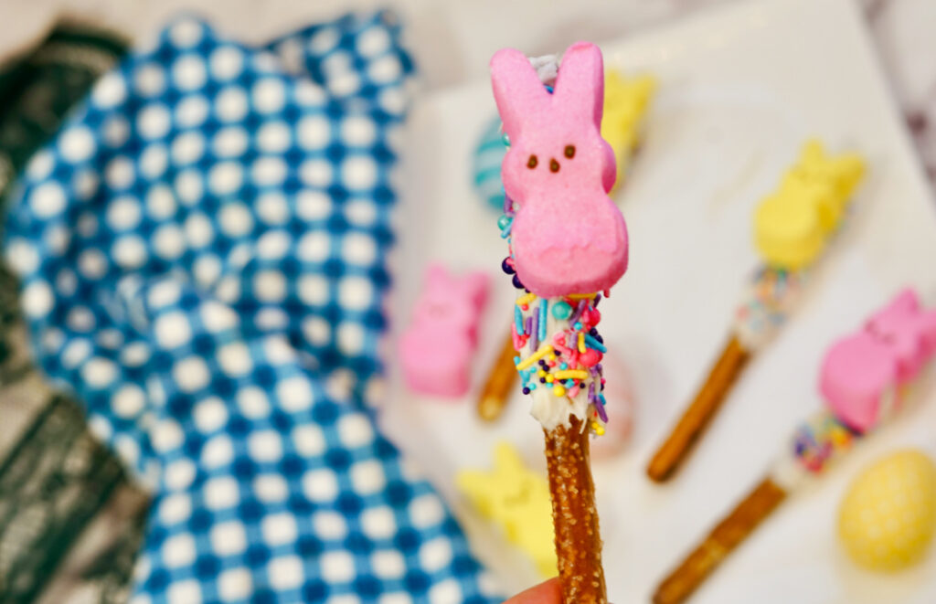 Peeps Pretzel Rods 6 A Peeps Pretzel Rod adorned with white icing, vibrant sprinkles, and a pink marshmallow bunny tops off the sweet treat. Similar decorated pretzels are scattered in the background on a blue and white-checkered cloth.