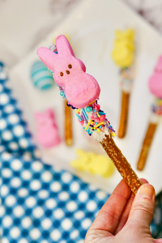Peeps Pretzel Rods 9 A hand holds a Peeps Pretzel Rod adorned with white chocolate, colorful sprinkles, and topped with a pink marshmallow bunny. In the background, other similarly decorated pretzel rods are arranged on a plate, each a festive treat in its own right.