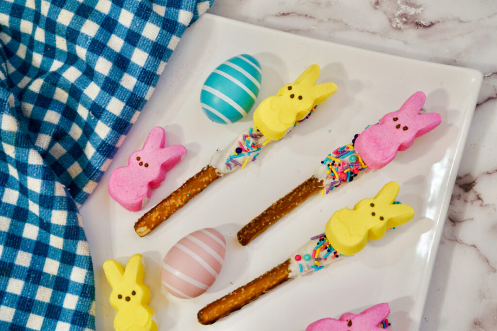 Peeps Pretzel Rods 2 Colorful marshmallow bunnies on Peeps Pretzel Rods with sprinkles, alongside decorated eggs on a white plate with a blue checkered cloth nearby. Peeps Pretzel Rods