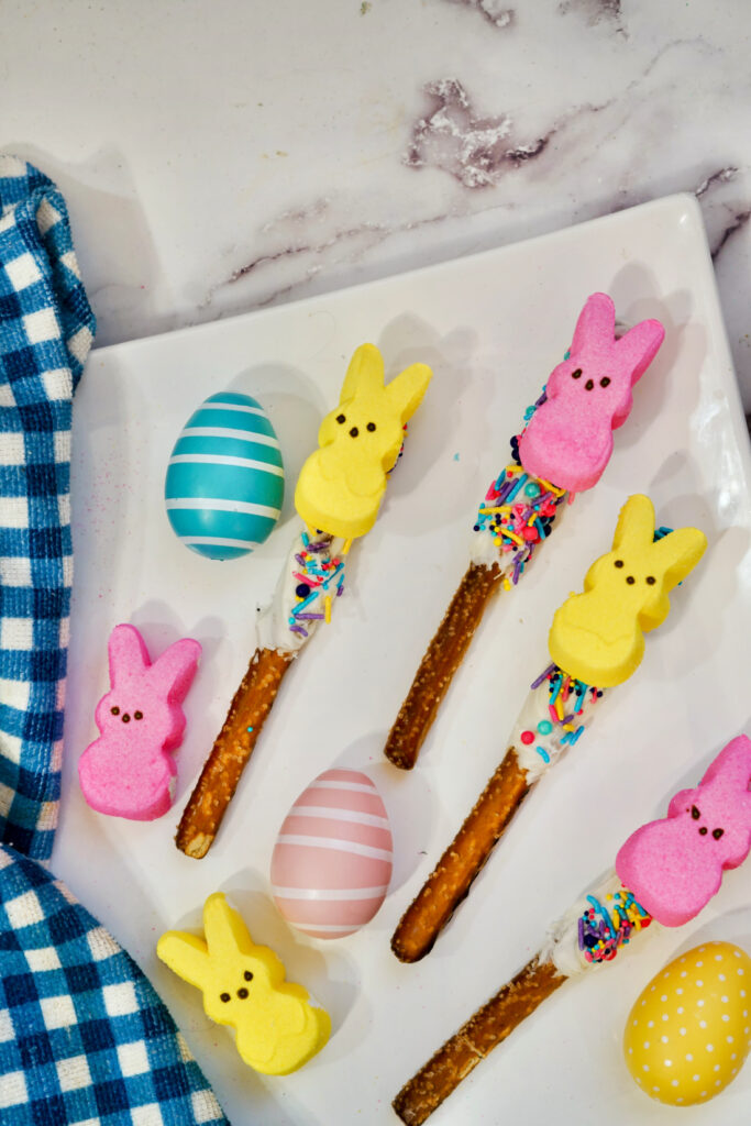 Peeps Pretzel Rods 7 Peeps Pretzel Rods adorned with marshmallow bunnies and sprinkles rest on chocolate-dipped sticks, accompanied by striped and polka-dotted eggs on a white plate with a blue checkered cloth alongside.