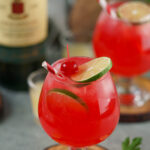 Two Irish Redhead Cocktails are adorned with lime slices and cherries, perched on wooden coasters. In the background, a bottle and extra garnishes complete the inviting scene.