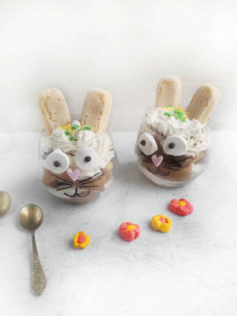 Tiramisu Easter Bunny – A Fun Twist on the Classic Italian Dessert 10 Two bunny-themed desserts in glasses, resembling playful Easter bunnies, come with whipped cream and ladyfingers as ears. They are decorated with candy eyes and flowers, all presented on a marble surface beside two spoons.