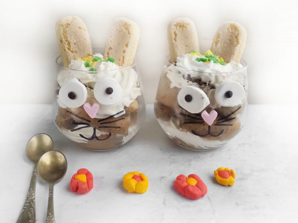 Tiramisu Easter Bunny – A Fun Twist on the Classic Italian Dessert 2 Two dessert glasses reminiscent of Easter bunnies feature bunny faces made from cookies, whipped cream, and candy. Each glass has biscuit ears like a playful twist on traditional tiramisu. Three small flower candies and two spoons complete the delightful scene.