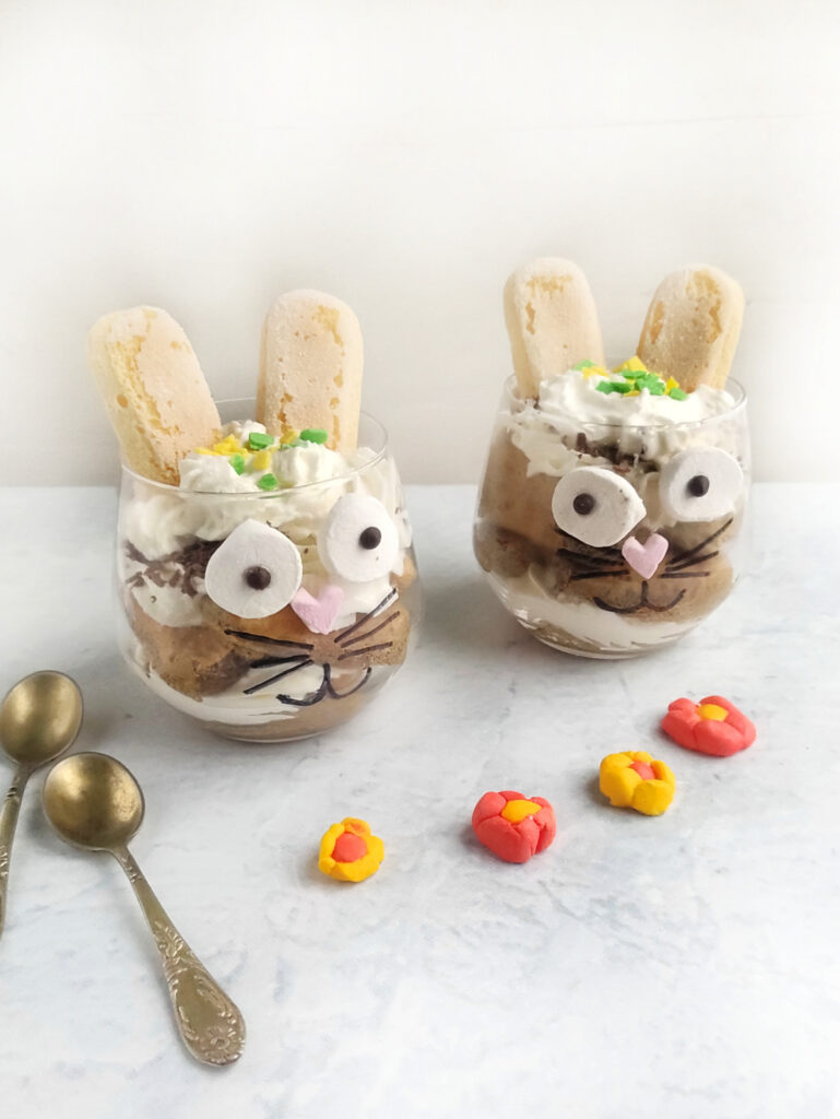 Tiramisu Easter Bunny – A Fun Twist on the Classic Italian Dessert 9 Two dessert cups resembling Easter bunnies are adorned with whipped cream, ladyfingers, candy eyes, and chocolate details. The display includes two spoons and small decorative flowers on a light surface—a whimsical tiramisu treat for the holiday.