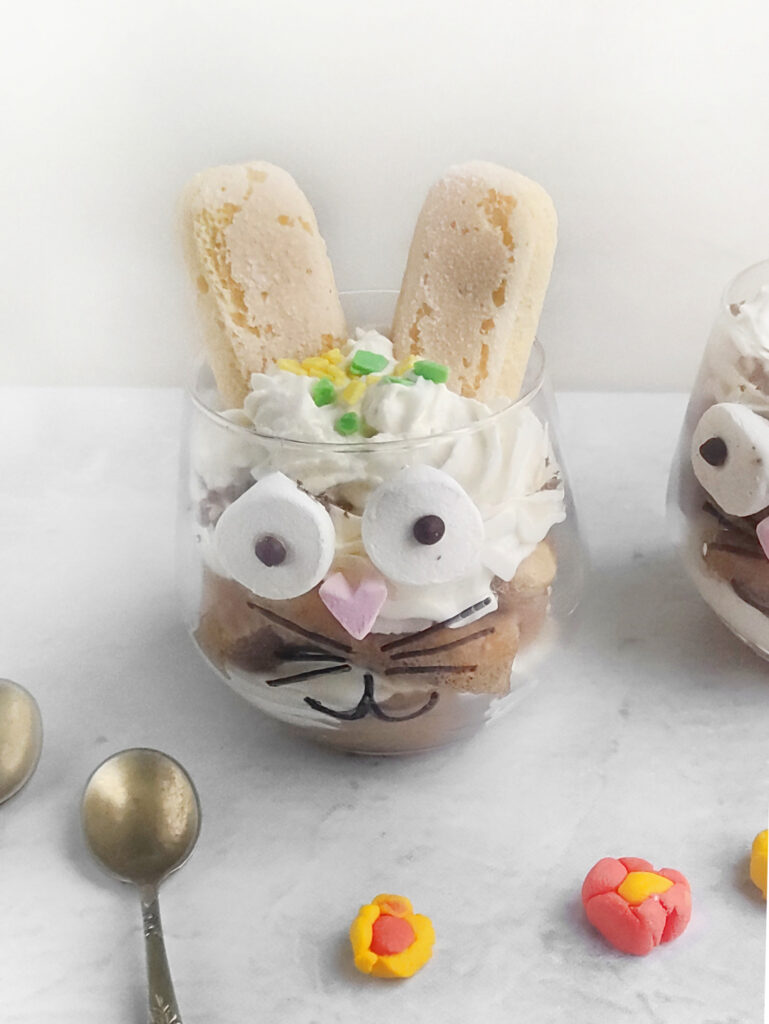 Tiramisu Easter Bunny – A Fun Twist on the Classic Italian Dessert 11 A glass filled with dessert, decorated to resemble an Easter Bunny tiramisu with cookies as ears, marshmallow eyes, and a heart-shaped nose. Two spoons and small candy flowers are nearby.