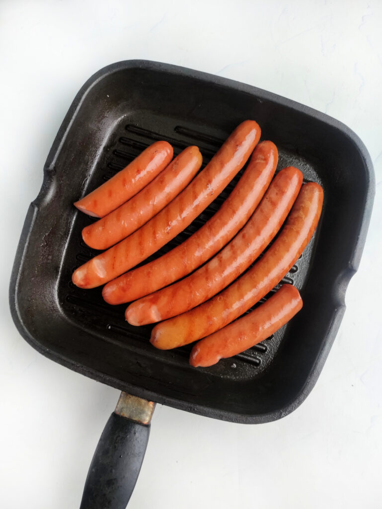 Hot Dog Patriot Celebration Board Recipe 4 Seven sausages are arranged in a grill pan, ready to be cooked on a white surface—perfect for your next hot dog celebration.