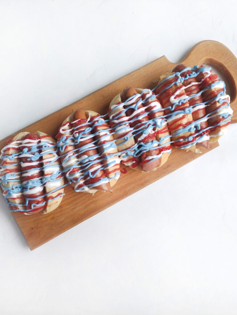 Hot Dog Patriot Celebration Board Recipe 7 A Hot Dog Patriot Celebration: four hot dogs on a wooden board, topped with red, white, and blue sauces, arranged in a festive row against a white background.