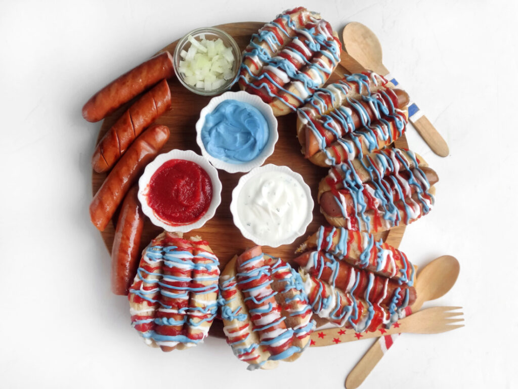 Hot Dog Patriot Celebration Board Recipe 2 A round Hot Dog Patriot Celebration Board featuring hot dogs with red, white, and blue sauce, sausages, bowls of chopped onions, ketchup, blue and white sauces, plus wooden cutlery and napkins—perfect for a festive gathering.