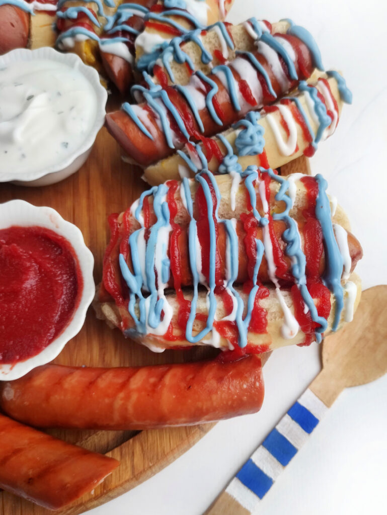 Hot Dog Patriot Celebration Board Recipe 10 Hot dogs take center stage on a Patriot Celebration Board, topped with ketchup and blue and white sauces. Served with bowls of dipping sauce, sausage pieces, and a wooden fork for the perfect festive bite.