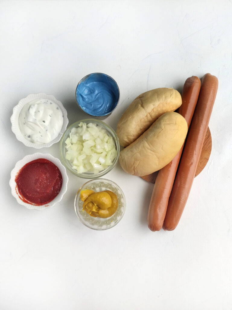 Hot Dog Patriot Celebration Board Recipe 3 Two hot dog buns and sausages are paired with small bowls of diced onions, ketchup, mustard, blue sauce, and white sauce on a white background—perfect for your Hot Dog Patriot Celebration Board.