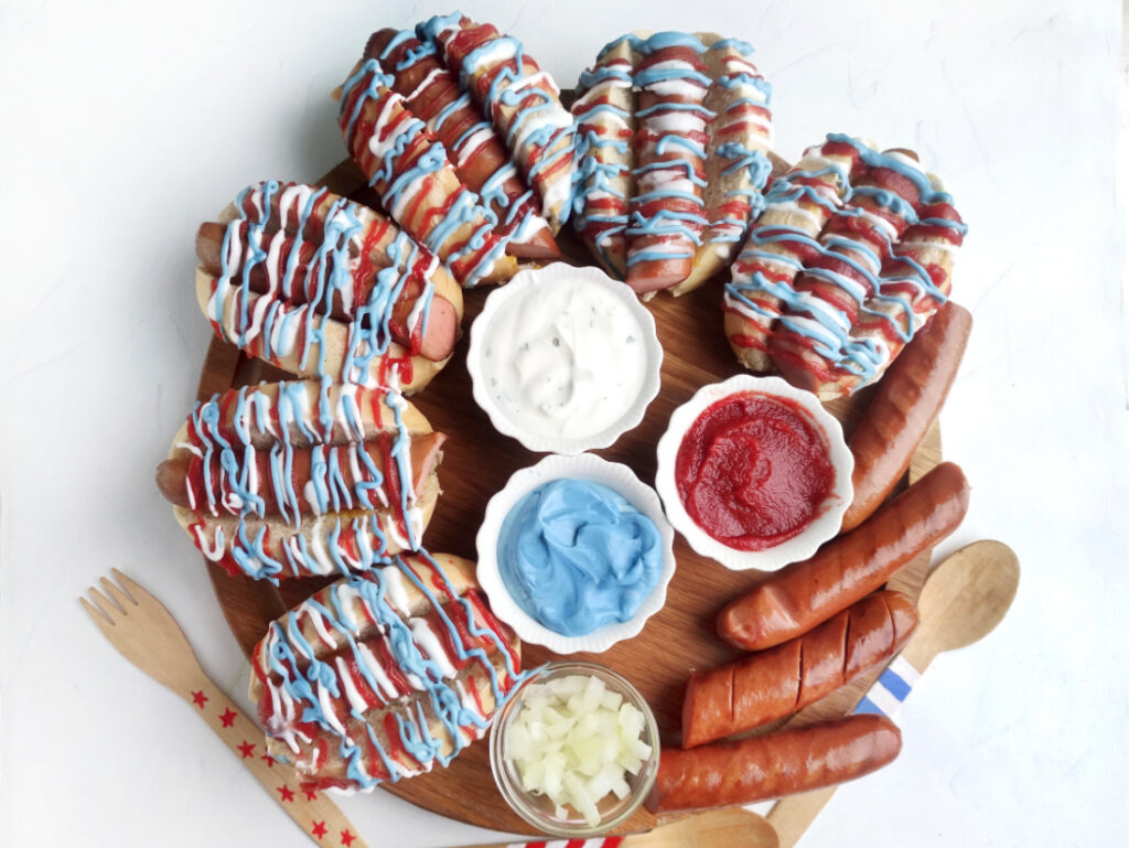 Hot Dog Patriot Celebration Board Recipe 9 A round Hot Dog Patriot Celebration Board featuring grilled hot dogs topped with festive red, white, and blue sauce, served with ketchup, ranch, blue dip, and chopped onions. Wooden utensils are on the side for easy serving.