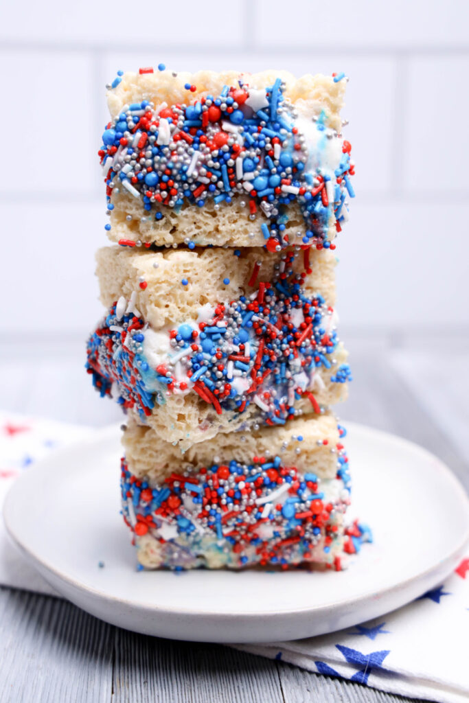 July 4th Rice Krispies Ice Cream Sandwiches 10 Three No-Bake Patriotic rice crispy treats, topped with red, white, and blue sprinkles, are stacked on a white plate against a light background.