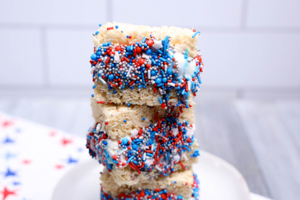 July 4th Rice Krispies Ice Cream Sandwiches 2 Three No-Bake Patriotic rice crispy treats stacked on a plate, partially dipped in white chocolate and covered with red, white, and blue sprinkles.