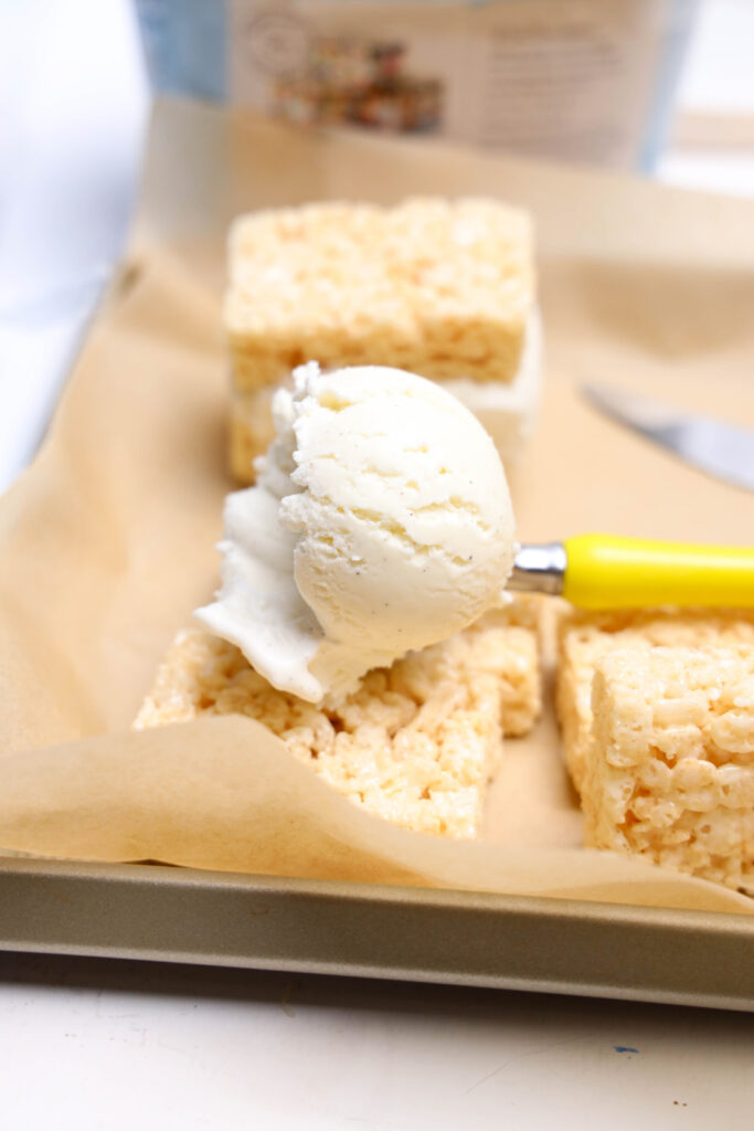 July 4th Rice Krispies Ice Cream Sandwiches 4 A scoop of vanilla ice cream sits on top of a rice krispie treat, ready to be made into Rice Krispies Ice Cream Sandwiches, with more treats and a yellow-handled ice cream scooper nearby.