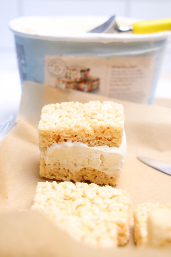 July 4th Rice Krispies Ice Cream Sandwiches 5 A close-up of Rice Krispies Ice Cream Sandwiches made with two rice crispy treats and vanilla ice cream on parchment paper, with a knife and ice cream container in the background—a perfect no-bake patriotic July 4th treat.