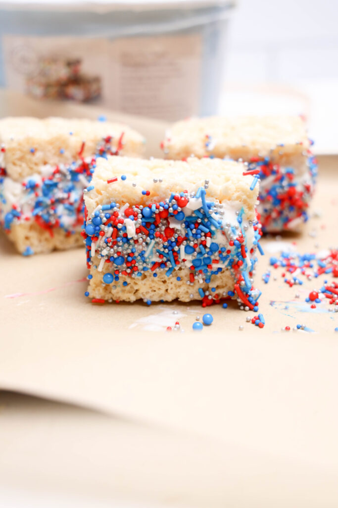 July 4th Rice Krispies Ice Cream Sandwiches 6 A No-Bake Patriotic Rice Krispies treat square with white icing and red, white, and blue sprinkles sits on parchment paper, surrounded by more festive treats—perfect for celebrating July 4th!.