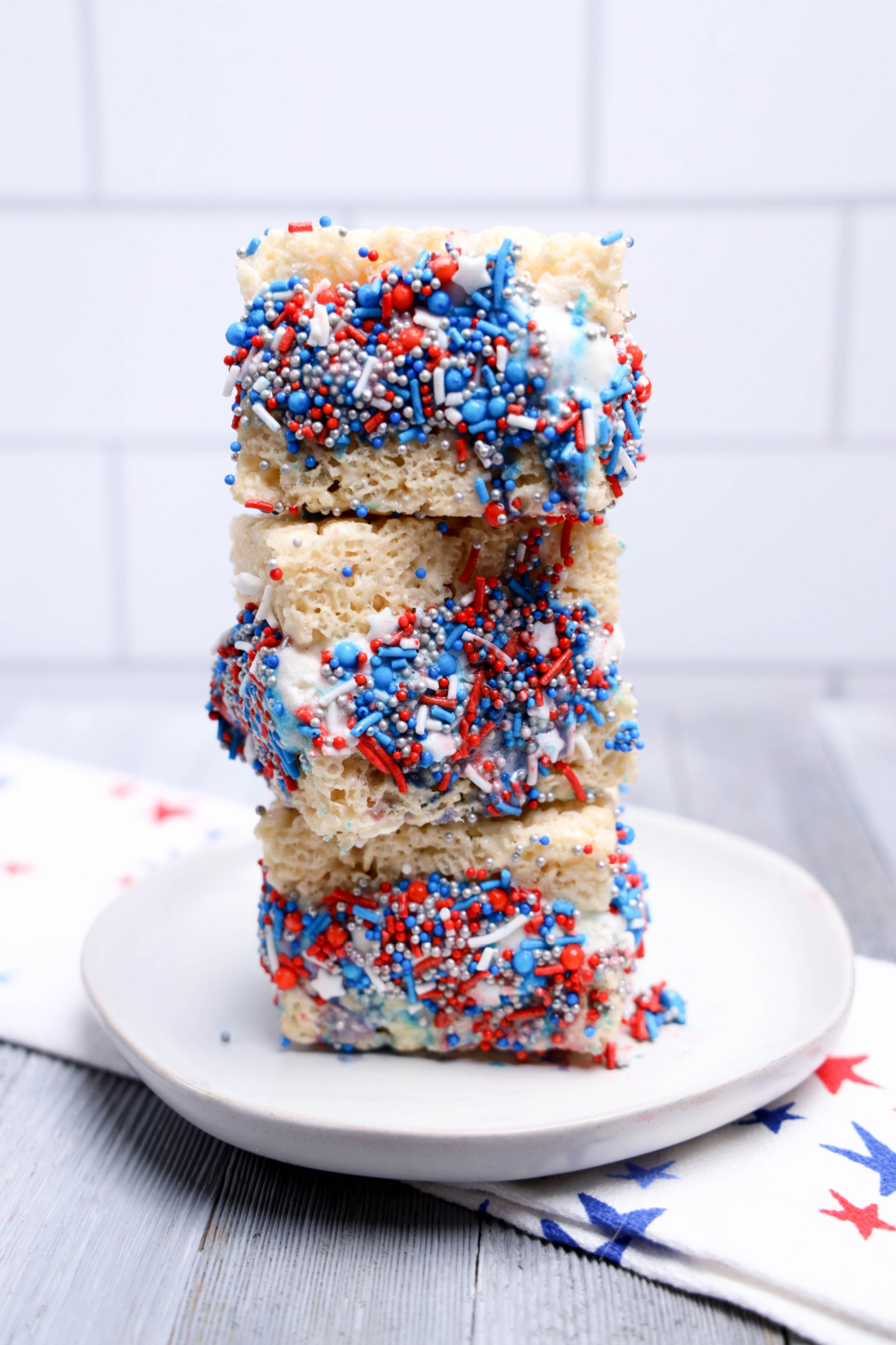 Three No-Bake Patriotic Rice Krispies treats stacked on a white plate, decorated with red, white, and blue sprinkles and icing, with a star-patterned napkin in the background.
